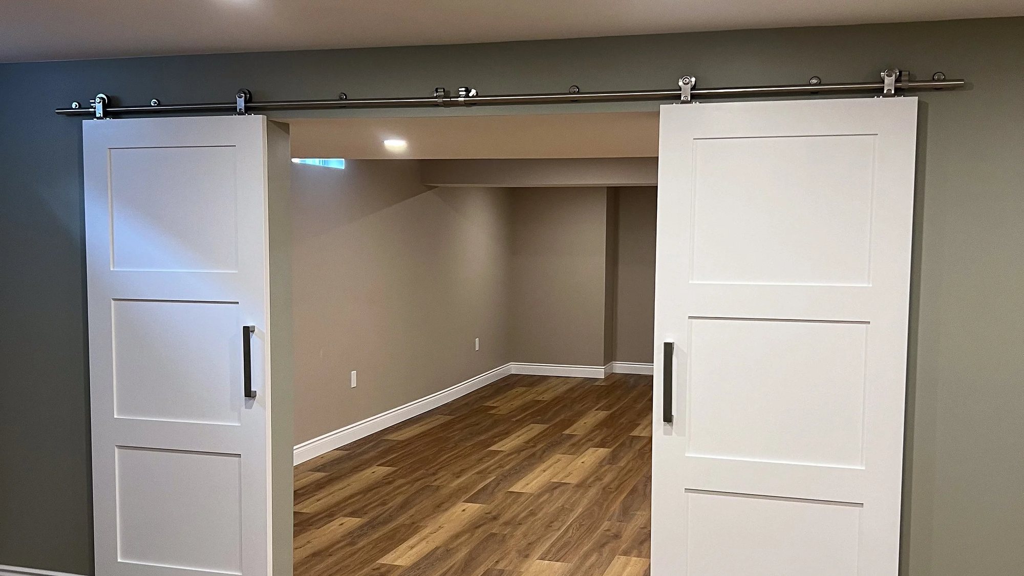 Finished basement with two white barn doors on a steel track, sage accent wall, wood-look plank flooring and recessed pot lights