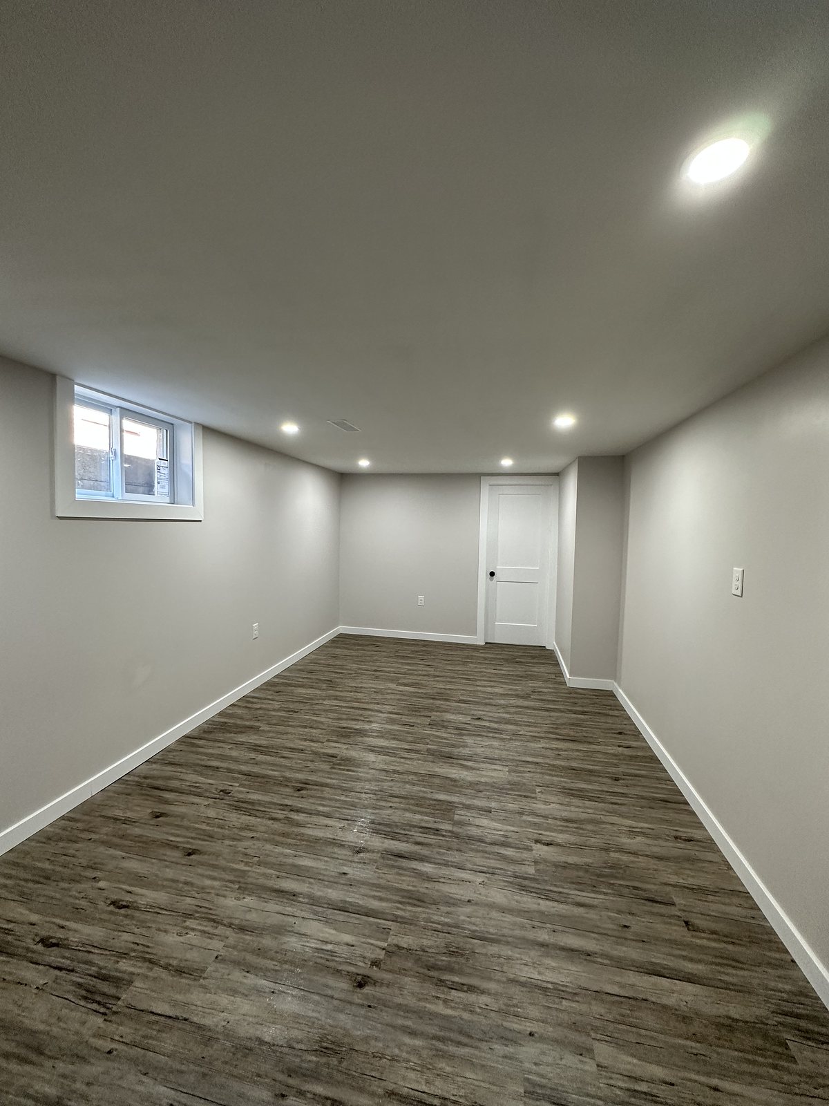 Finished basement with grey walls, wood-look plank flooring, pot lights and an egress window