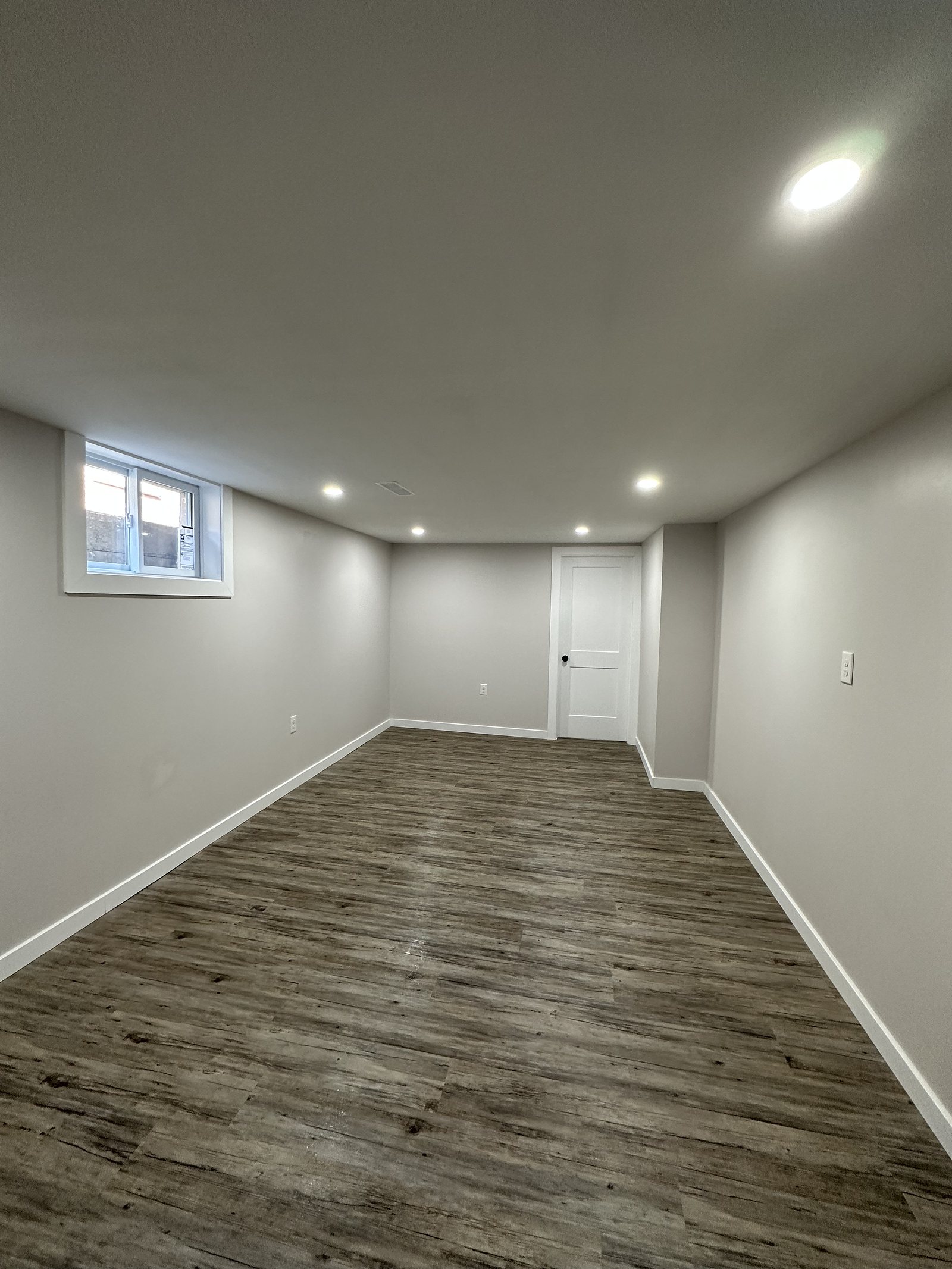 Finished basement with grey walls, wood-look plank flooring, recessed pot lights and an egress window