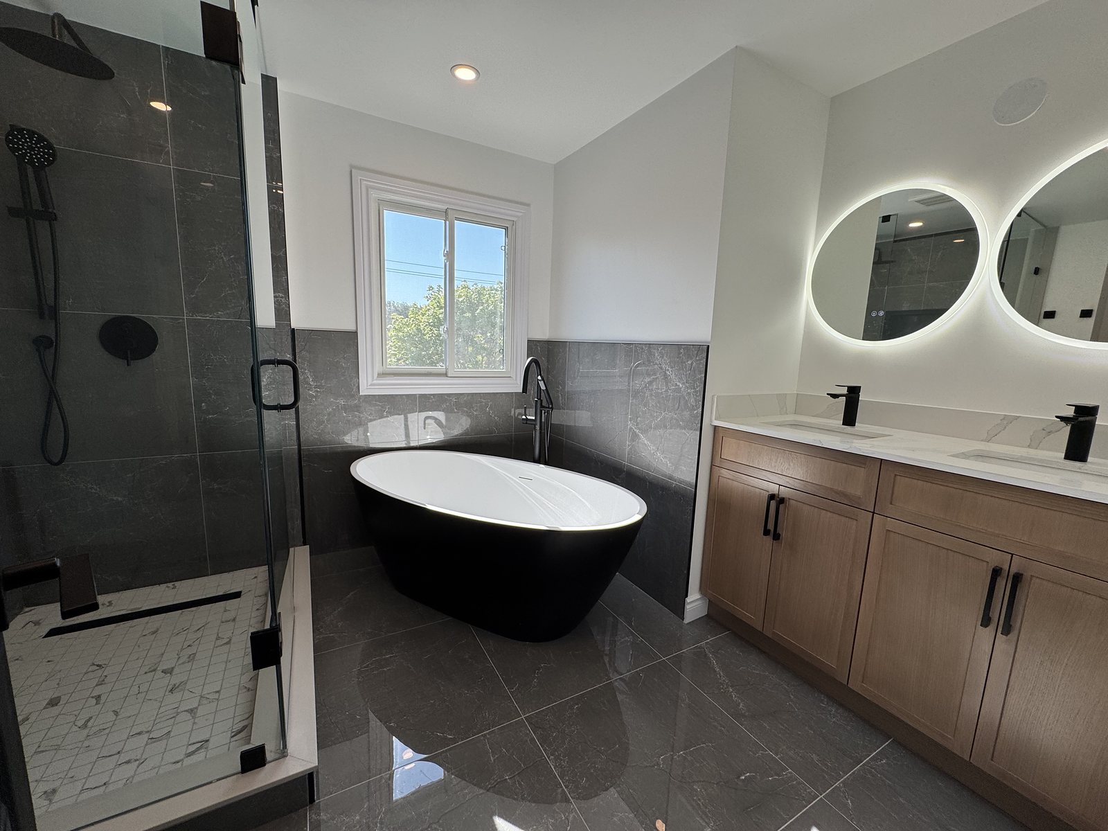 Double vanity with backlit round mirrors beside a freestanding tub